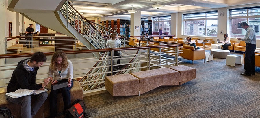 The Pardee Management Library buzzing with patrons, some working together and others on laptops at desks, with stacks and the staircase behind